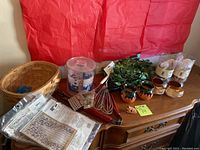 Photo showing a wooden dresser top with a woven basket, three southwestern style ceramic mugs, artificial green plant, BBQ utensils, three glass candles, and two curtain packages.