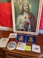 Framed religious picture of Jesus Christ with heart, placed on wooden dresser along with other religious items including medallions, booklets, plaques, and crosses.