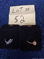 Two rings displayed on black velvet pads with lot number 52 shown on a handwritten paper. One ring is silver-tone with rhinestones arranged in a leaf or feather pattern, the other is gold-tone with a floral cluster design of clear stones.