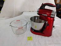 Red Betty Crocker electric stand mixer with stainless steel mixing bowl and clear Pyrex measuring cup with red markings beside it.