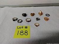 Photo of 10 assorted rings laid out on white cloth with yellow lot number card. Rings vary by color, size, and design including stones and engravings. One black rubber band included.