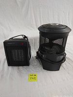 Photo showing two floor heaters side by side on a white cloth background. The left heater is a black rectangular Homestyles heater with visible mesh grill and power cord. The right heater is a round, black Garrison heater with mesh body and control knob on top, cords wrapped around base.