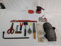 Wide view of 13 assorted hand and measuring tools laid out on white cloth including a red 24" Kapro level, red scissors, a black mallet, and other tools along with knee pads.