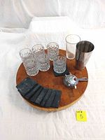 Full set overview on wooden round cutting board, including 6 whiskey glasses, cocktail shaker, cocktail strainer, and 8 slate coasters.