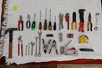 Photo showing all 28 items from the tool bundle laid out on a white cloth, including hammer, pliers, wrenches, clamps, screwdrivers, drill bits, staple gun, measuring tape, and other hand tools.