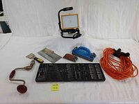 View of eight tools including LED work light, hand crank drill, wall scraper, concrete trowel, rasp, hand sander, extension cord, and drill bit set