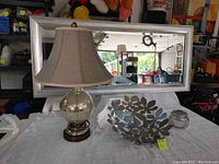 Image of vanity set showing large silver painted framed mirror, bubble lamp with beige fabric shade, silver metal decorative flower bowl, and small silver candle holder jar.