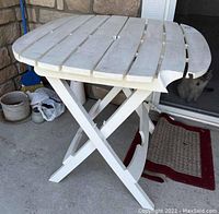 Side angle of the white folding round plastic patio table showing slatted top, folding legs, and chip on corner.