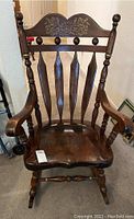 Front view of dark wood rocking chair with carved and painted floral design on top of headrest, spindle backrest, armrests, and visible scratches on seat.