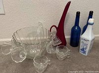 Image showing clear glass punch bowl with scalloped edges, multiple clear glass cups with flower-shaped handles surrounding the bowl, two blue glass bottles, white and blue decorative bottle, and tall red glass vase.