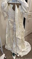 Front and side views of vintage ivory wedding dress on hanger showing puffed sleeves, floral brocade fabric, lace neckline, and flowing skirt with train.