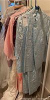 Photo showing several vintage women's dresses and ball gowns hanging on hangers including a greenish-blue floral brocade two-piece suit with large buttons visible on the jacket.
