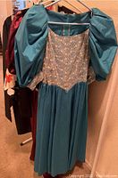 Photo showing teal vintage dress with puffed sleeves, lace bodice, and flowing skirt on hanger along with two additional dresses on rack.