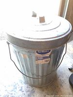 Photo showing galvanized steel trash can labeled NorWestCo with handle and lid on concrete floor.