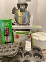 Full view of the lot showing the waffle maker, boxed Magic Bullet blender, Farberware vegetable ricer, and multiple mini muffin pans stacked in the foreground.