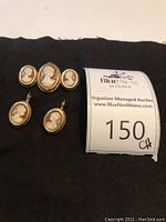 Full set on black cloth background including brooch with three cameos and matching pair of earrings, next to auction lot tag