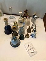 Photo showing an assortment of 13 collectible bells of various types and materials arranged on a white surface with a lot tag numbered 108 visible.