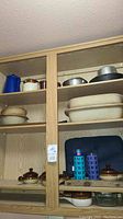 View of kitchenware on wooden shelves including Pampered Chef stoneware pots and crocks, blue enamel coffee pot, metal pans, Pyrex casserole dishes, and water bottles.