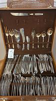 View of the open wooden flatware case with bronze flatware pieces arranged inside, showcasing forks, knives, and spoons of various sizes, some wrapped in plastic.