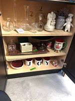 Cabinet shelf showing various glass vases, jars, holiday mugs and plates spelling 'JOY', and small decor items.