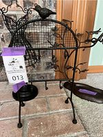 Tall black metal bird-themed jewelry holder with multiple hooks, standing on floor near brick wall and wooden door