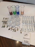 Full lot view including four colored drinking glasses, eight glass salt cellars with spoons, twelve glass cutting utensil rests, and six wine charms arranged on white surface.