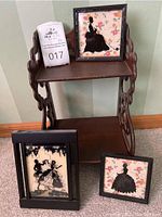 Photo showing the wooden shelf with two silhouette paintings displayed on the floor and one on the top shelf, along with auction ticket 017