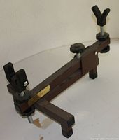 Photo shows a brown metal adjustable shooting rest with a horizontal bar supported by two vertical legs. At each end of the horizontal bar are padded v-shaped supports with adjustment knobs. The frame shows minor signs of light use.