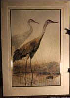 Framed fine art print showing two sandhill cranes standing in a marshy area with dry grasses and water, matted in cream-colored custom matting, soft lighting with slight shadow on right edge
