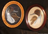 Two oval duck portraits, one in light wood frame and one in dark wood frame, both with bubble glass covering the paintings showing side profile duck pairs.