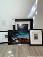 Four framed art pieces arranged on floor showing black frames and varied sizes including metal print center front