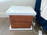 Photo showing one bedside table with white glossy top and base, wood grain drawer front, against light wall and floor