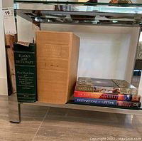 Seven books displayed on glass shelf, including a large beige dictionary and green law dictionary, along with titles about the Vatican and travel.