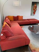 L-shaped red upholstered sofa with striped and solid color pillows in red, orange, and mustard tones, shown from an angled side view with ottoman and floor lamp behind.