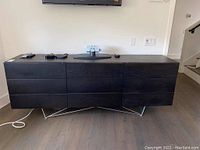 Front view of dark wood credenza cabinet with drawers and metal legs, placed against white wall with TV mounted above it.