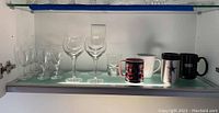 Seven clear wine glasses, two clear shot glasses, and five assorted mugs on glass shelf inside cabinet.