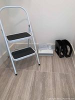 White metal step ladder with two black non-slip steps, two black plastic magazine holders, and two white metal desk organizers with cutout design arranged on floor.