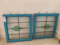 Two antique stained glass panels in distressed blue wooden frames leaning against wall showing same design motif each 22x44 inches