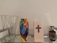 Four vases against wall: tall clear glass, colorful geometric ceramic, rectangular ceramic with black cross, small Asian-style ceramic with crackled glaze and painted figures