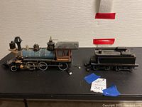Two-piece model train including a locomotive and coal car on a black table against a light textured wall.