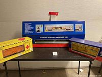Three train car boxes: USA Trains passenger car in blue box center flanked by yellow and purple boxcars packaging on table