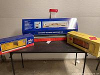 Three boxed G scale train cars: USA Trains extruded aluminum passenger car in original blue box, USA Trains Union Pacific boxcar in yellow box, Aristo-Craft boxcar in gold box.