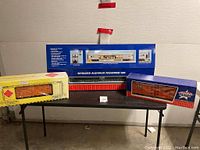Three boxed model train cars displayed on table: passenger car in blue box, Aristo-Craft boxcar in yellow box, USA Trains Scout boxcar in red/blue box