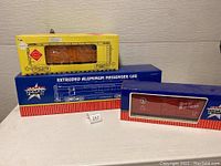 Three boxed 1:29 scale model train cars shown stacked: USA Trains blue box with passenger car, Great Northern red boxcar, yellow Aristo Craft reefer car box.