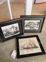 Three framed black and white drawings arranged on the floor showing two buildings and one sailing ship.