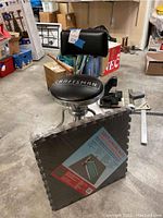 Craftsman workbench chair with black padded seat, backrest, and metal frame, shown alongside packaged soft lining mats on concrete floor.