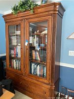 Front view of extra-large wooden cabinet with two glass doors showing shelves filled with books and decor items, cabinet rests on carpeted floor near blue wall.