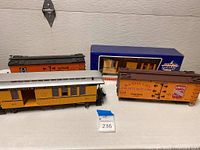 Four G scale train cars arranged on white surface showing Denver & Rio Grande Western passenger car, Kansas City beef line refrigerated boxcar, Santa Fe boxcar, and scrap car.