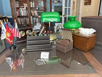 Green banker desk lamp, green glass oil lamp, wooden tissue holder, brown leather penholder organizer, coasters, and trashcan on glass table with bookshelves in background.