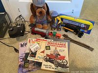 Wide view showing plush dog toys in engineer outfits, boxed yellow model train, curved train tracks, tanker truck toy, wooden train car, and several magazines.